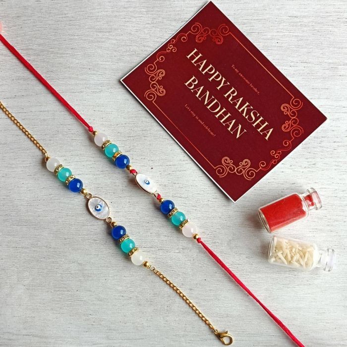 metal charm evil eye rakhi set for bhaiya bhabhi placed on textured sheet with roli chawal bottle and rakshabandhan card
